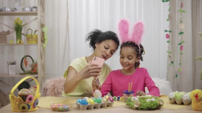 妈妈和女儿带着有趣的兔子耳朵用电话在视频通话中交谈。非裔美国妇女和小女孩坐在装饰房间的桌子旁。复活节