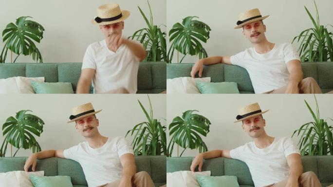 现代创意时尚潮人的肖像戴着小胡子，戴着帽子，圆形粉色太阳镜坐在沙发上，在现代阁楼公寓里眨眨眼。千禧一