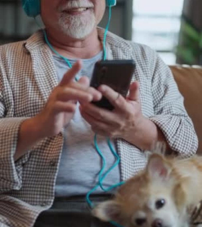 亚洲老人戴着耳机，戴着狗最好的朋友宠物坐在沙发上，一边听音乐一边唱歌，一边在家舒适的客厅休息