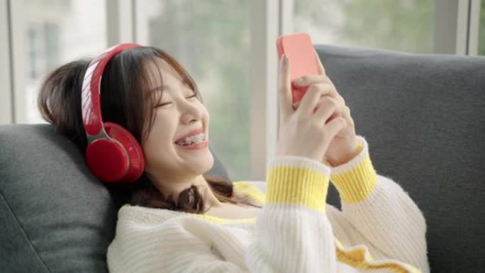亚洲少女躺在沙发上，用耳机听音乐，用智能手机在社交媒体、休闲概念、社交距离、物联网上交流