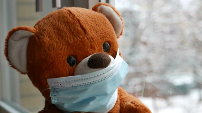 好奇的泰迪熊，戴着新型冠状病毒肺炎的面罩，检疫透过窗户看着户外飘雪