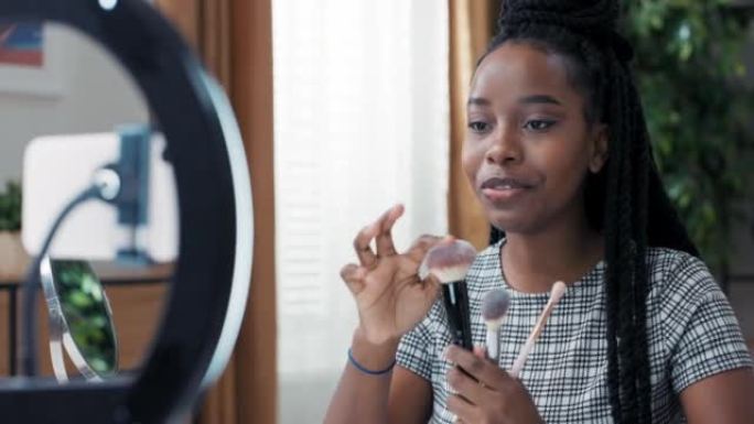 一个黑皮肤的女人，辫子扎在马尾辫上，录制了有关如何选择合适的化妆刷的简短教学视频。女孩在社交媒体上创