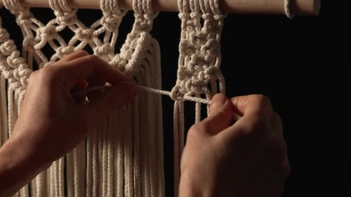 女人用自己的手编织花边线的图案，用于装饰或家居装饰。在黑色背景的创意工作坊中，女人的手在绳子上打结。