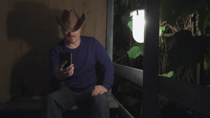 牛仔男子坐在门廊上用手机在线交流。夜晚
