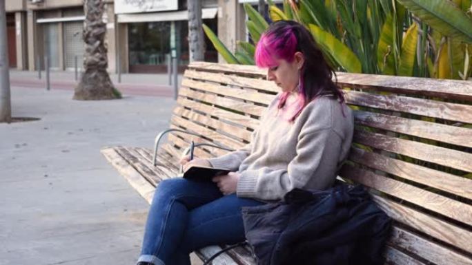 粉红色头发的女人坐在公园长椅上的笔记本上写字
