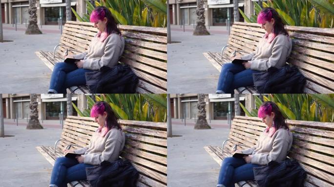 粉红色头发的女人坐在公园长椅上的笔记本上写字