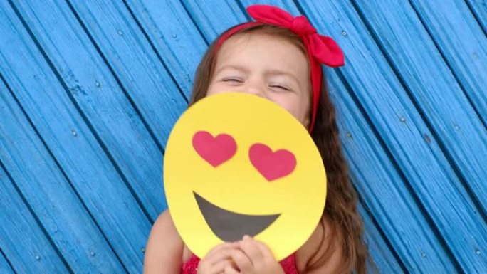 可爱的黑发小女孩用一颗心的纸板笑脸遮住她的脸。世界表情符号日。情感爱。
