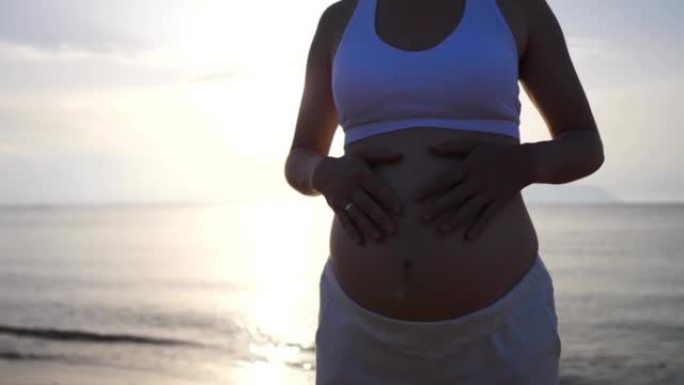 在柔和的夕阳下，海滩上孕妇的剪影。婴儿出生前的女人会花时间在新鲜空气中。一个女人抚摸着她的大肚子，里