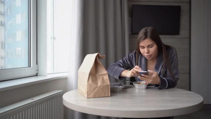 女孩坐在家里，用一次性铝箔饭盒在餐桌上吃外卖的食物。使用智能手机滚动浏览社交网络的新闻。惊喜的情感。