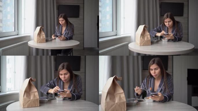 女孩坐在家里，用一次性铝箔饭盒在餐桌上吃外卖的食物。使用智能手机滚动浏览社交网络的新闻。惊喜的情感。