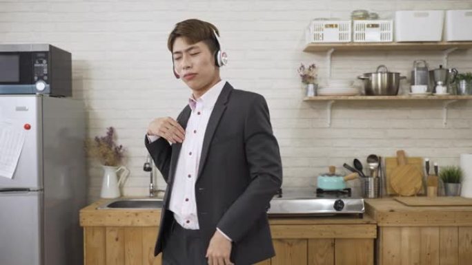 慢动作炒作的亚洲商人戴着耳机，一边用食指指着，一边在家里的厨房里随着音乐跳舞。