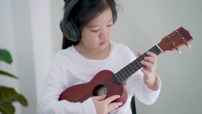 小可爱5岁初级亚洲女孩戴着无线耳机听音乐，跟随音乐节奏演奏尤克里里。音乐教室。乐器。弦乐器。