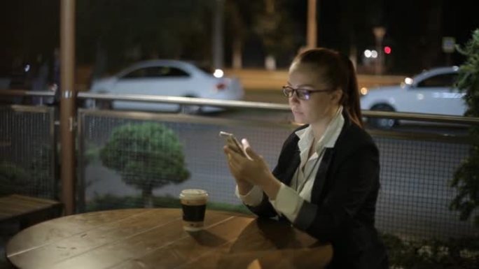 美丽的年轻白人妇女晚上下班后坐在街头咖啡馆喝咖啡，并使用智能手机在社交网络上聊天。