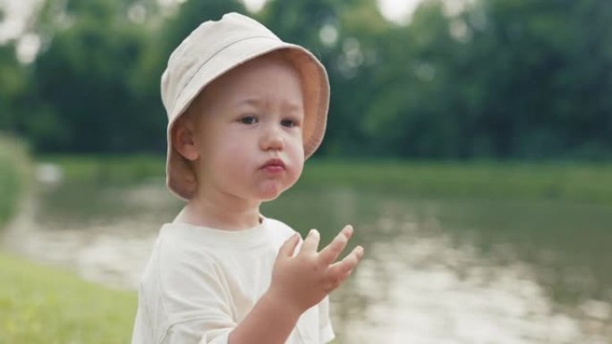一个戴着帽子，正在吃水果的小孩脸上的特写镜头，男孩坐在靠近水，湖的草地上，一个有爱心的父亲用食物擦拭