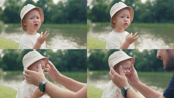 一个戴着帽子，正在吃水果的小孩脸上的特写镜头，男孩坐在靠近水，湖的草地上，一个有爱心的父亲用食物擦拭
