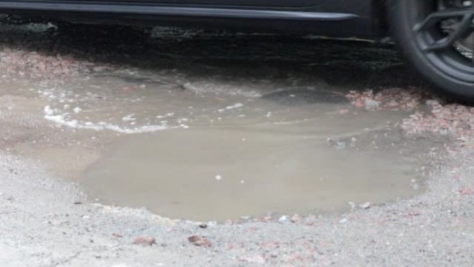 有过往汽车的柏油路面上充满水的坑洼。多雨的天气。白天下雨后，汽车在破碎的柏油路面上穿过水坑。雨滴和汽
