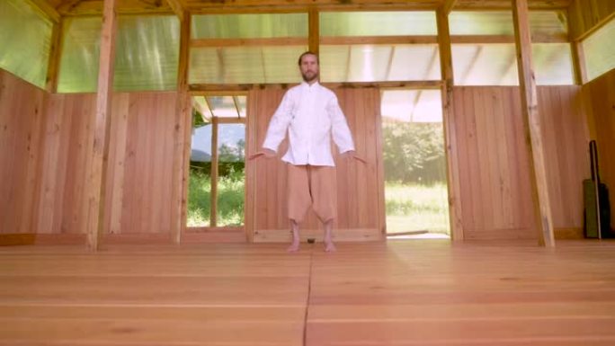 一个白种人留着金色长发的大胡子男人在夏季森林的木制练习室练习气功和太极拳。欧洲领土上的武术练习