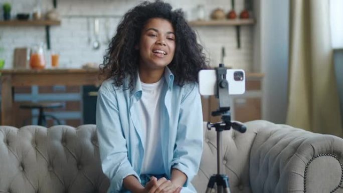 年轻的非洲裔美国女性博客作者vlogger坐在家里的沙发上录制视频或在智能手机上直播。博客和视频广播
