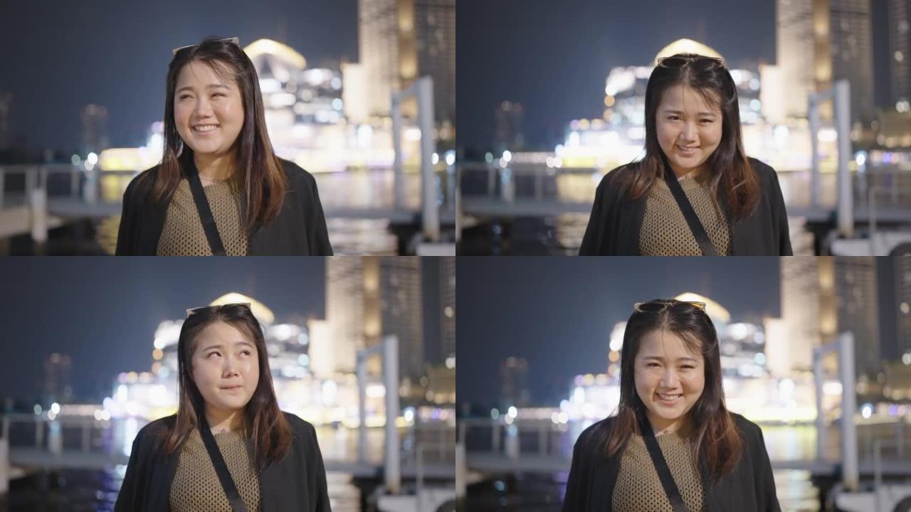 亚洲妇女微笑着看着相机，而快乐地站在户外的城市现代城的夜晚。