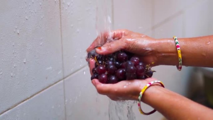 印度女性双手的特写镜头在家里水龙头的水流下小心地清洗黑葡萄果实。洗水果和蔬菜，保健。