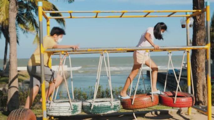 戴着口罩的亚洲女孩和她的兄弟在户外操场上玩耍，带着阳光，活跃和新的正常生活方式理念。