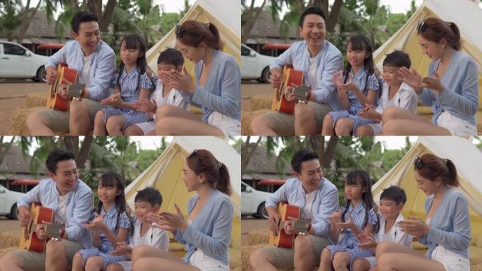 快乐的亚洲家庭坐在帐篷前，边露营边弹吉他