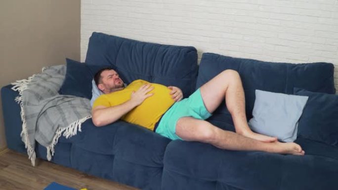 穿着黄色t恤的胡子丰满的男人躺在家里的床上，抚摸着生病的大肚子。腹胀，暴饮暴食，痉挛和疼痛，腹泻。适