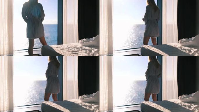 一个穿着浴袍的和平快乐的年轻金发女郎，在阳光明媚的日子里，她的早晨咖啡站在全景窗户的背景下，俯瞰大海