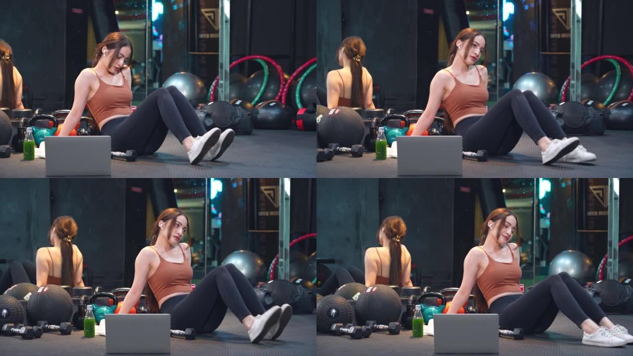 一名亚洲女性在健身房，在健身在线小时用笔记本电脑锻炼后正在休息