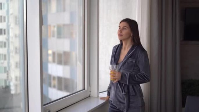 人们和度假概念快乐的高加索女人早上穿着休闲服在家站在窗户附近喝柑橘汁或冰沙。