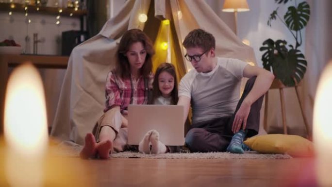 母亲，父亲和可爱的孩子坐在玩具wigwam附近，使用笔记本电脑