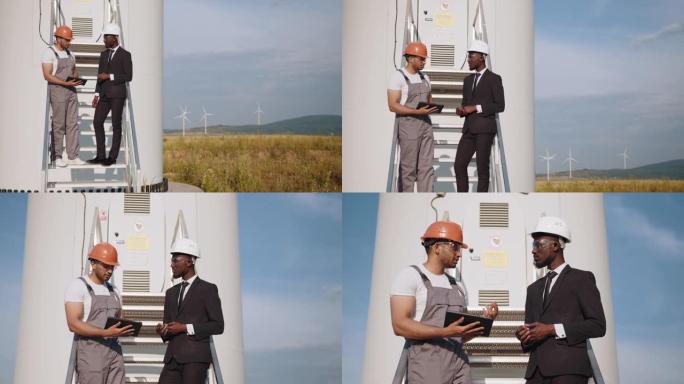 穿着橙色头盔和灰色工作服的印度工程师指着远处，拿着平板电脑靠近风车。穿着西装的非裔美国商人在检查农场