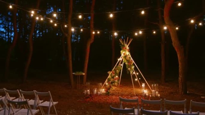 晚上松林户外婚礼场地波西米亚提皮拱门装饰上的节日串灯照明。夏季乡村婚礼上，复古串灯灯泡花环闪耀在椅子