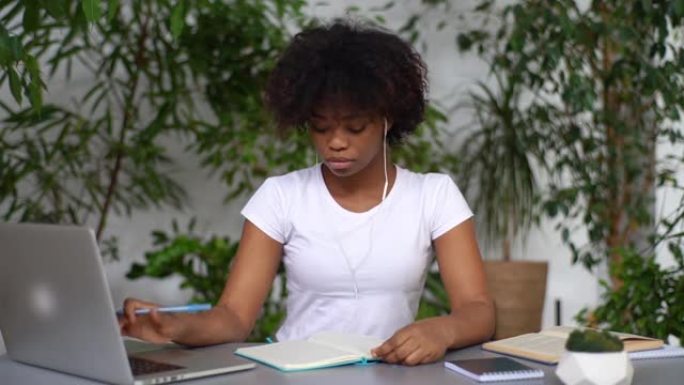 迷人的非裔美国年轻女子坐在桌子上，使用笔记本电脑，通过耳机收听音频课程，在字帖上做笔记