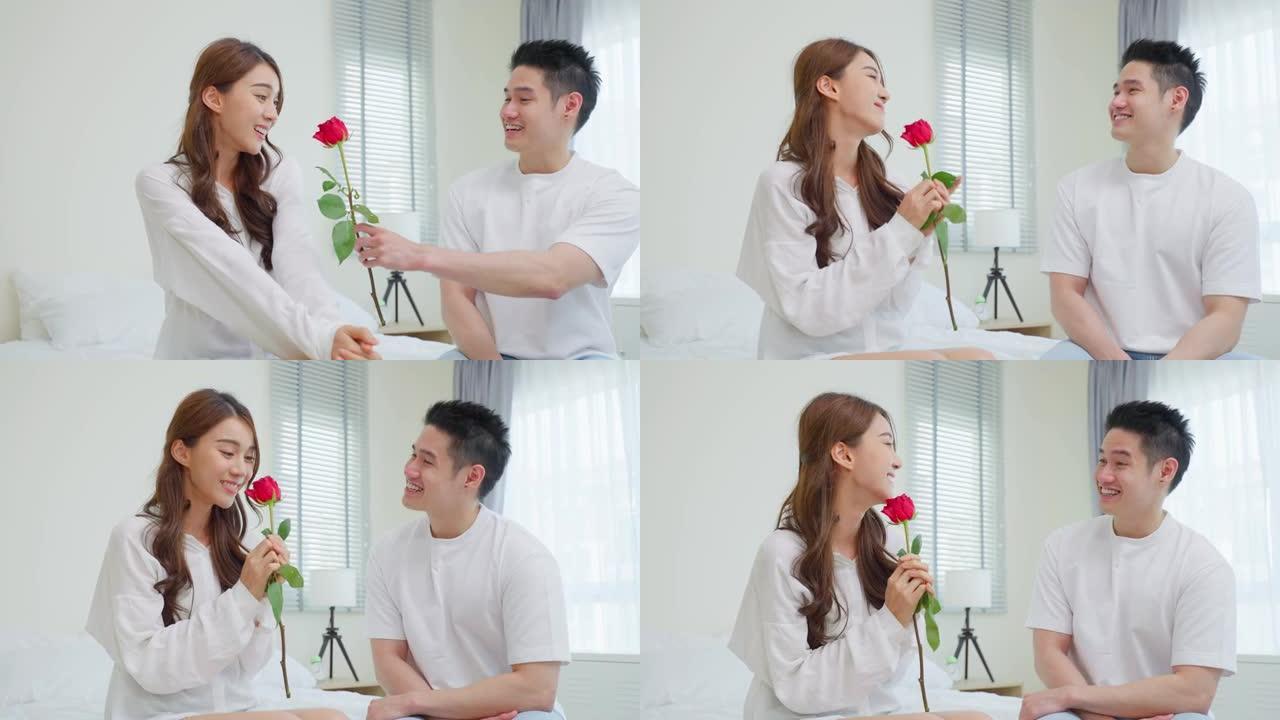 亚洲年轻人惊喜并将玫瑰花送给美丽的女友。迷人的浪漫新人男女坐在床上，一起庆祝周年纪念日和情人节