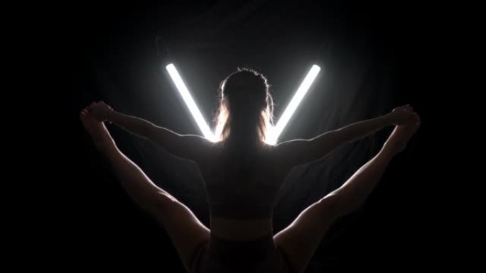练习瑜伽姿势的健康女性。剪影女孩在黑色背景下的工作室里做运动，带v形白色发光二极管灯管。无压力内部平
