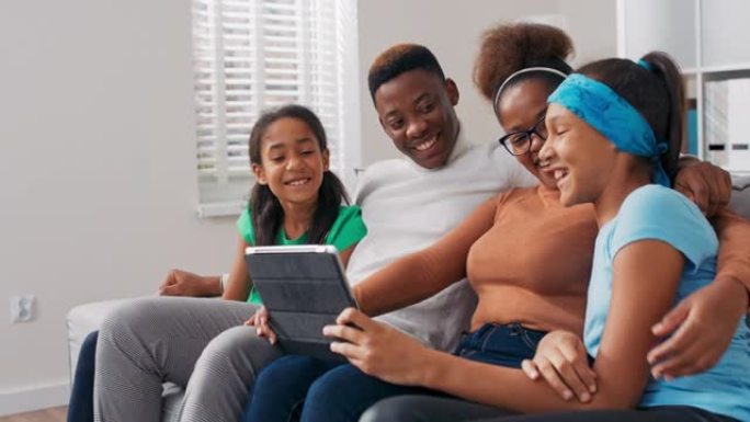 一个幸福的家庭坐在沙发上，放松身心，下午一起度过时光，父母带着两个女儿在平板电脑上挥手，与朋友进行视
