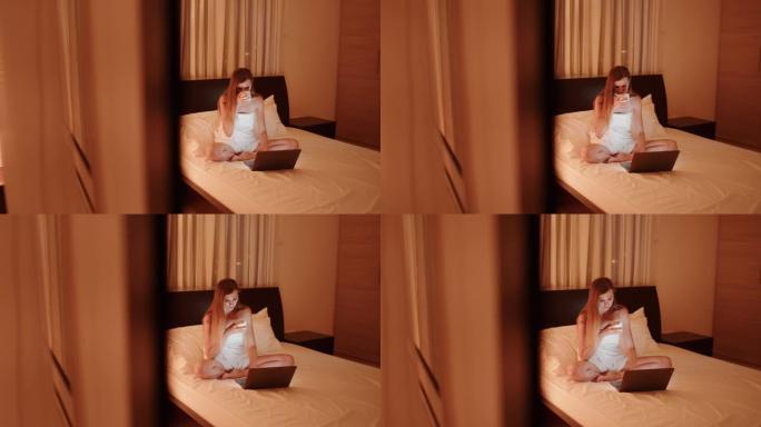 疲倦的女人晚上在床上使用笔记本电脑。晚上坐在酒店房间床上的喜出望外的女士。穿着浴巾的漂亮女人用笔记本