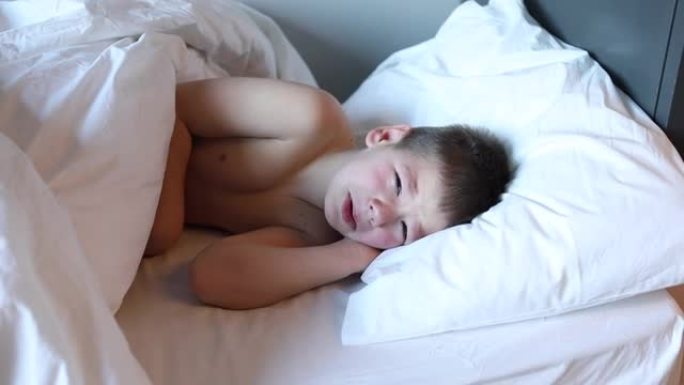 昏昏欲睡的蹒跚学步的男孩躺在床上，穿着白色床上的衣服哭泣。卧室里累了的孩子。