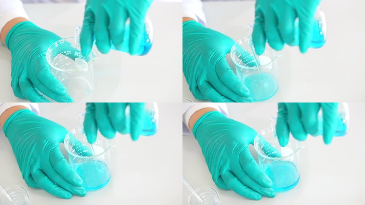 专业化学家的特写手穿着防护制服，戴着手套，将移液器中的溶液加入到烧瓶中的蓝色混合物中。在现代实验室工