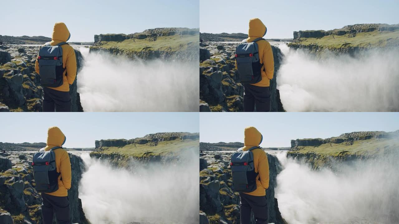 穿着黄色夹克和背包的男子站在悬崖边看着冰岛的Detifoss瀑布享受