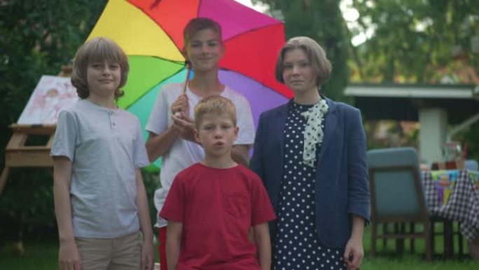 中等镜头正面的高加索男孩和女孩看着相机微笑着旋转彩色雨伞站在户外。快乐的朋友在春夏后院慢动作摆姿势。