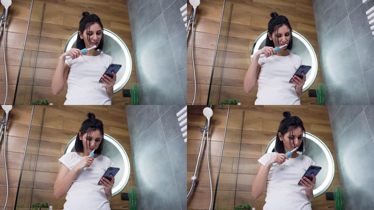 近距离观察满意的漂亮黑发年轻女子，她站在浴室的镜子旁，在刷牙时使用手机