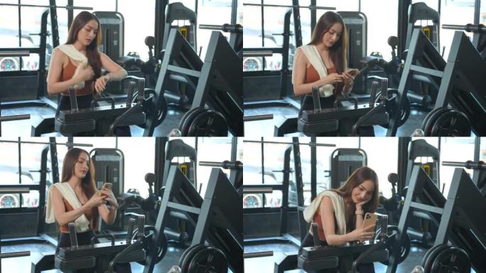一名亚洲妇女在健身房休息，在健身时间用减肥机锻炼后，用毛巾擦干脸，喝有机蔬菜汁，用智能手机放松。
