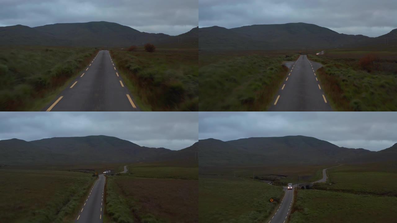 乡村狭窄道路上方的低空飞行。用拖车提升并揭示迎面而来的越野汽车。背景中的山脊。爱尔兰