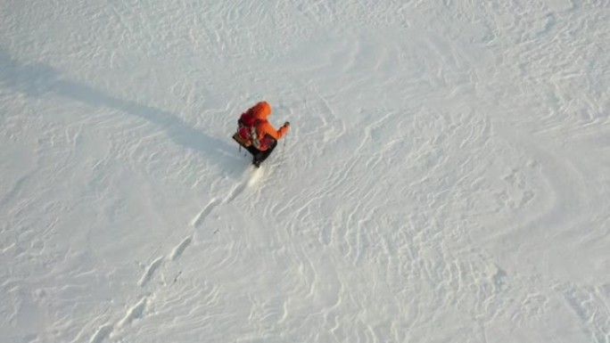 空中4k镜头-一个背着背包和棍棒的人走过白雪皑皑的沙漠，留下了一连串的脚印。在极冷条件下生存的概念