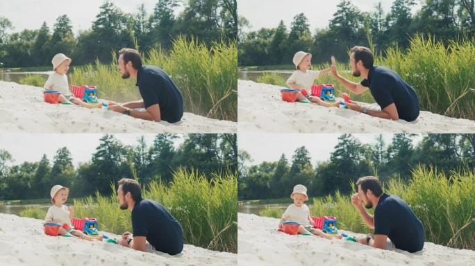 一位父亲跑向一个戴着帽子的小男孩，在海滩上的一个湖边玩耍，躺在儿子旁边的沙滩上，和他说话，他们击掌，