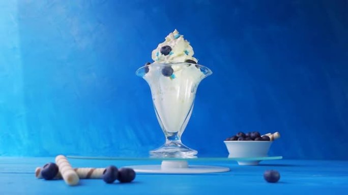 蓝色背景的木制桌子上的托盘上的玻璃旋转蓝莓冰淇淋。具有速度斜坡效果的创意慢动作4k循环视频。