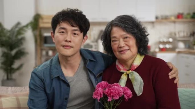 微笑的亚洲母亲和成年儿子在家里的客厅里看着相机，手里拿着一束康乃馨。世代爱情与母亲节概念