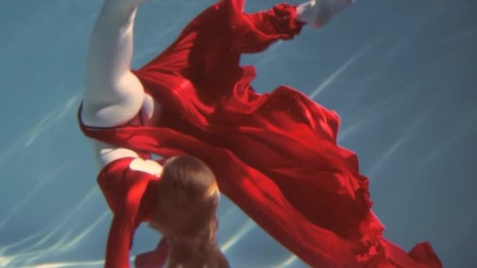 迷人的红发年轻女子穿着红色连衣裙美丽地在水下游泳。慢动作